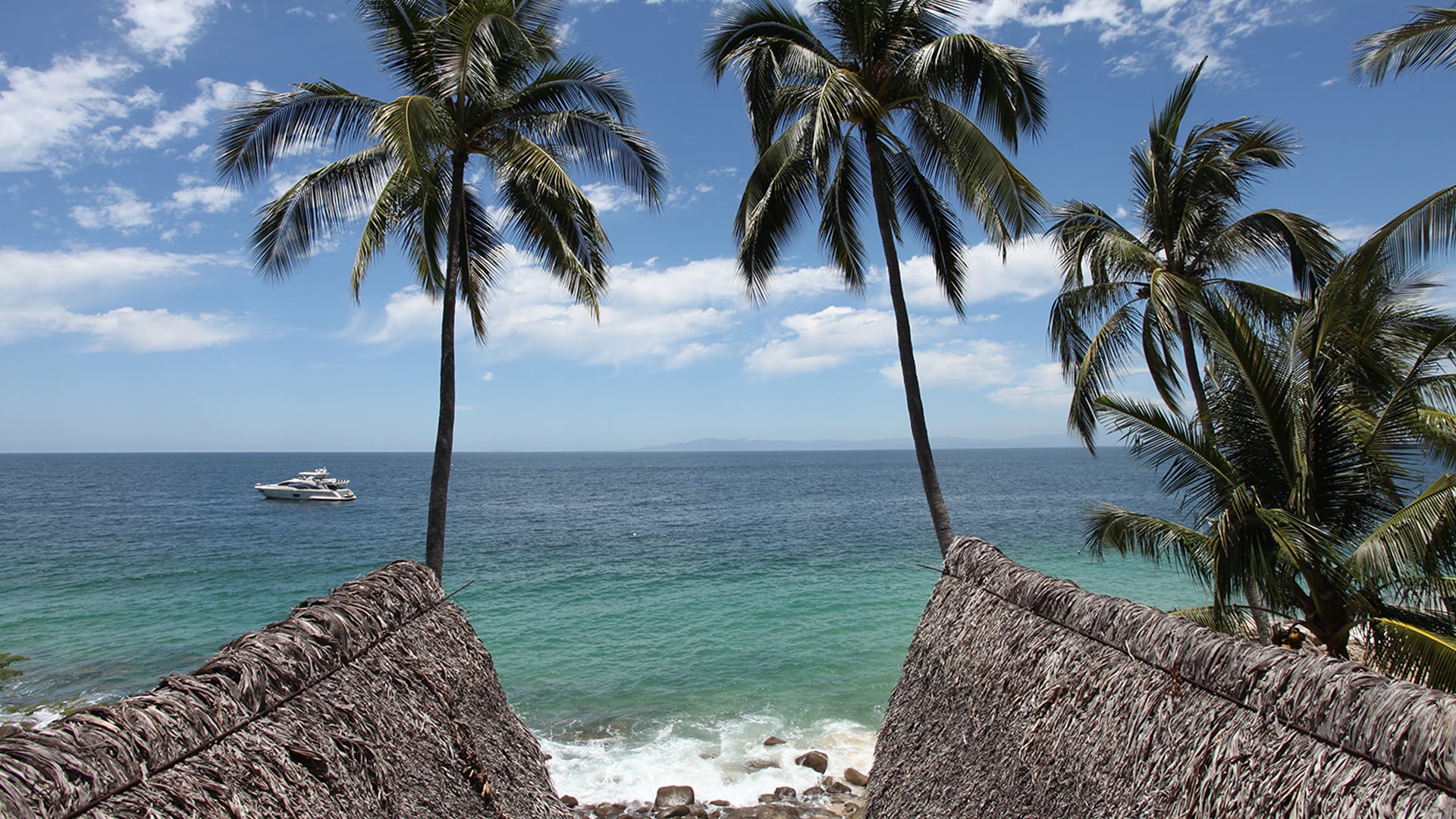 Two palapas on the beach with palm trees and a powerboat anchored nearby